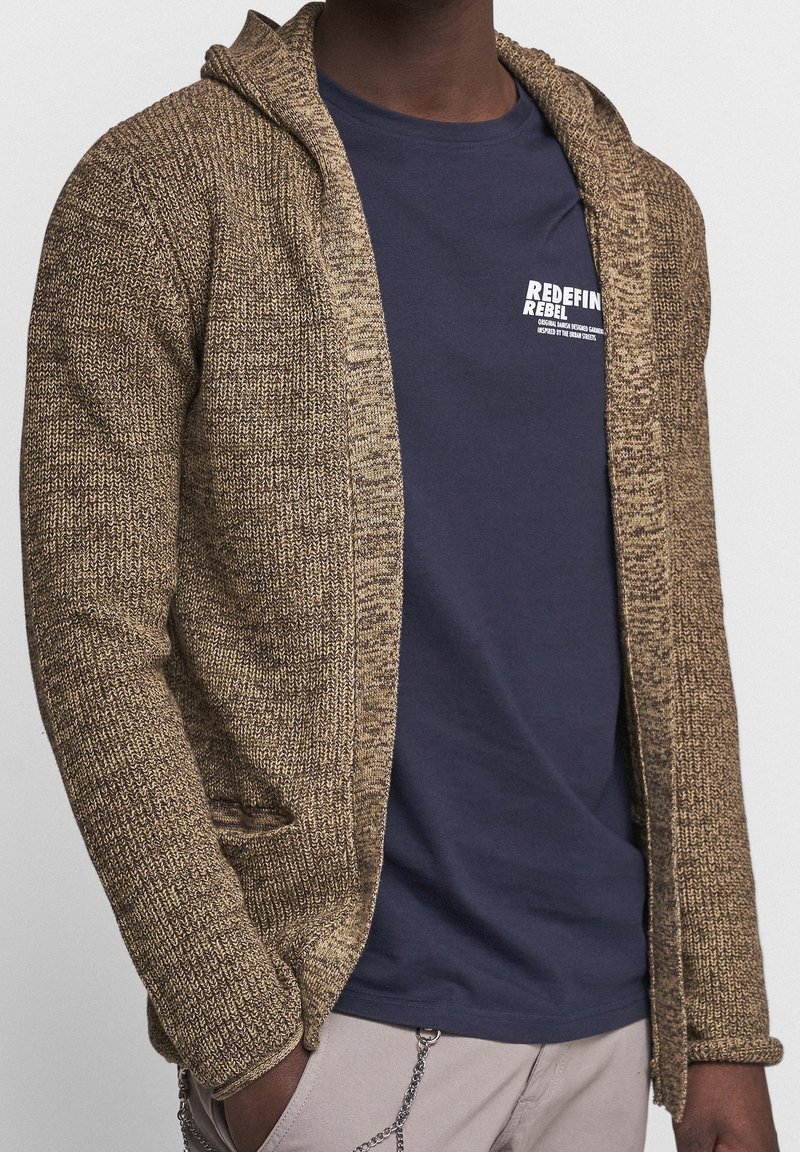 Personne portant un cardigan à capuche en maille marron par-dessus un t-shirt bleu marine avec un texte blanc, un pantalon beige et un accessoire chaîne à la taille.