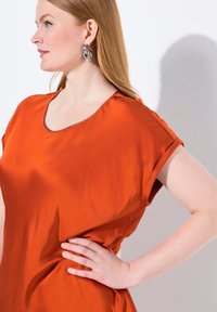 Top arancione in satin con maniche corte arrotolate, scollatura a girocollo e un leggero lucido. Il modello ha i capelli mossi e indossa un grande orecchino d'argento.