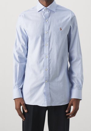 Homme portant une chemise bleu clair à fines rayures, manches longues, col boutonné et logo brodé sur la poitrine, associée à un pantalon noir.