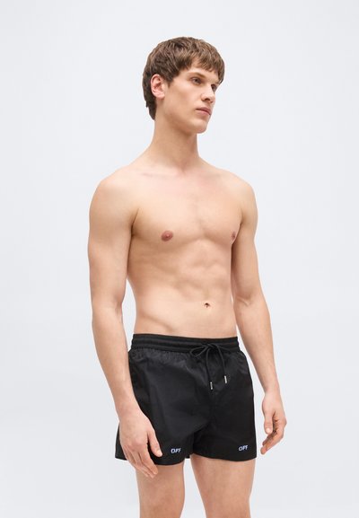 Jeune homme torse nu portant un short de bain noir avec le texte blanc « OFF » sur chaque jambe, debout devant un fond gris clair uni.