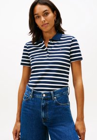 Nainen, joka pukeutunut laivastonsiniseen ja valkoiseen raidalliseen lyhythihaiseen poolopaitaan, joka on tungettu korkeavyötäröisiin sinisiin farkkuihin, seisoo yksinkertaisen taustan edessä.