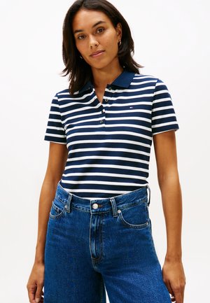 Naine, kes kannab tumesinise ja valge triibulise lühikeste varrukatega polosärki, mis on toppitud kõrge vöökohaga sinistesse teksadesse, seisab ühevärvilise tausta ees.