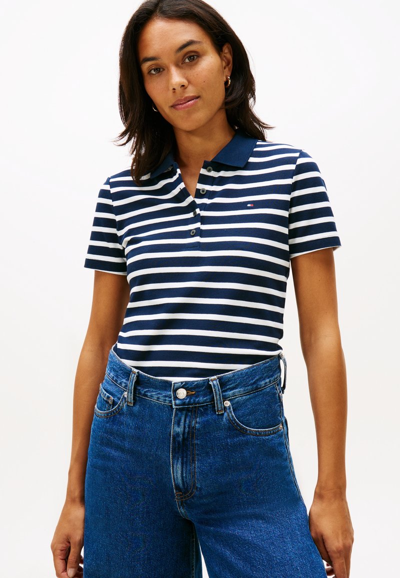 Nainen, joka pukeutunut laivastonsiniseen ja valkoiseen raidalliseen lyhythihaiseen poolopaitaan, joka on tungettu korkeavyötäröisiin sinisiin farkkuihin, seisoo yksinkertaisen taustan edessä.