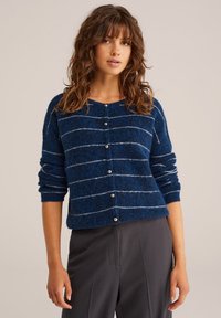 Modra prugasta cardigan s puhasto teksturo, z gumbi na sprednji strani in dolgimi rokavi. Ima kontrastne bele pruge in okrogle gumbe.