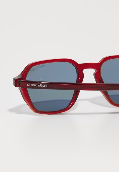 Červené plastové slnečné okuliare s ostrými rámami, modro zafarbenými sklami a metalickým doplnkom s nápisom "Giorgio Armani" na ramene.