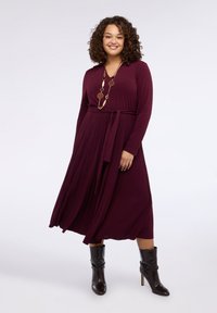 Bordowa sukienka z długim rękawem i dekoltem w serek, z paskiem w talii i rozkloszowanym dołem. Noszona z czarnymi botkami i naszyjnikiem w złotym odcieniu.