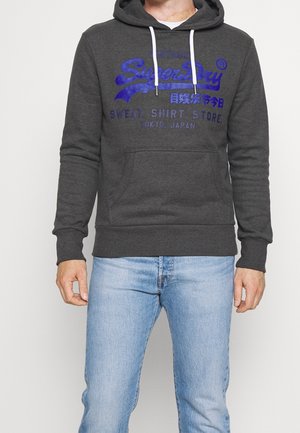 Mężczyzna w ciemnoszarej bluzie z kapturem z niebieskim napisem „SuperDry” i jasnoniebieskich dżinsach, stoi na tle gładkiego jasnego tła.