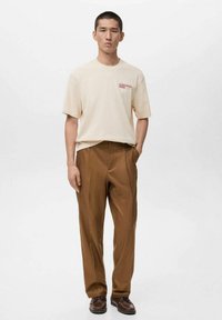 Beige cotton t-shirt with red text, paired with brown pleated trousers, and dark brown shoes, model stands against a plain light background.
