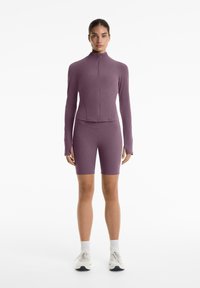 Lila Sportoutfit bestehend aus einem langeärmeligen Top mit Reißverschluss und Daumenlöchern sowie passenden Shorts. Hergestellt aus dehnbarem Stoff, der für Komfort sorgt.