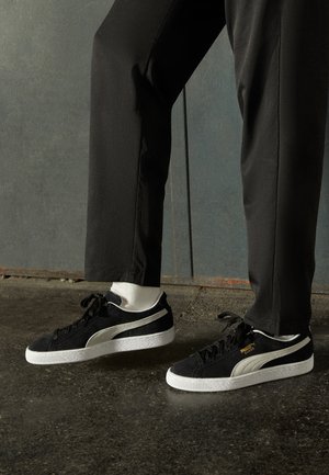 Personne portant un pantalon noir, des chaussettes blanches et des baskets Puma Suede noires debout sur un sol sombre près d'un mur gris.