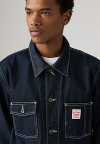 Mørkeblå denimjakke med krage, to frontlommer og gullknapper. Utsmykket med hvite sting og en liten rød merkeetikett.