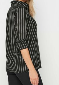 Schwarz- und grau gestreift, mit einem Kragen zum Knöpfen, hochgerollten Ärmeln und einem lockeren Schnitt. Das Shirt hat vertikale Streifen in unterschiedlicher Dicke.