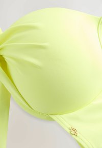Neon žuti bikini gornji dio od glatkog, rastezljivog materijala. Sadrži uvrnut dizajn sprijeda i mali zlatni detalj sa strane.