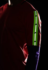 Czerwony sportowy T-shirt z zielonym i odblaskowym paskiem na ramieniu, z czarnymi logo Nike i kontrastującym niebieskim materiałem na rękawie.