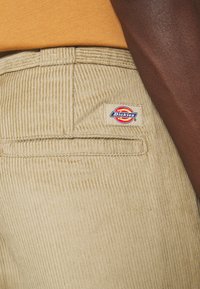 Beiga kordfloddsbyxor med vertikala ribbor, en bakficka och en Dickies logotags. Midjan syns något mot en brun yta.