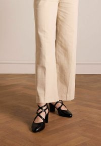 Scarpe con tacco alto in pelle nera con cinturini incrociati alla caviglia e punte affusolate, abbinate a pantaloni larghi beige chiaro su un pavimento di legno.