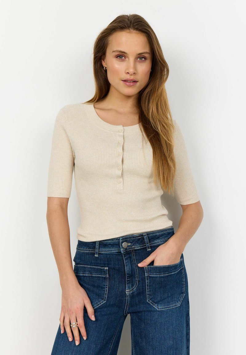 Ljusbeige ribbad t-shirt med kort ärm och rundad halsringning, prydd med fem knappar fram, kombinerad med mörkblå högmidjade jeans.