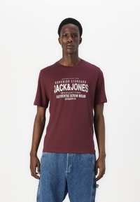 Burgundy cotton t-shirt featuring white text, round neck, and short sleeves. Worn with blue denim shorts, displayed against a white background.