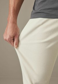 Ljusbeige, ribbade bomullshängselbyxor med en figursydd silhuett, hållna vid midjan av en hand. Tyget ser mjukt och elastiskt ut.