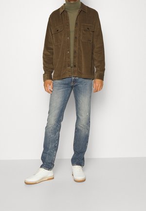 Homme portant une veste en velours côtelé marron, un col roulé vert olive, un jean bleu délavé et des baskets blanches, se tenant devant un fond blanc uni.