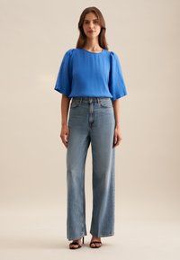 Top in lino blu con maniche corte a palloncino, abbinato a jeans a vita alta blu chiaro con gamba larga. Sandali neri con tacco completano l'outfit.