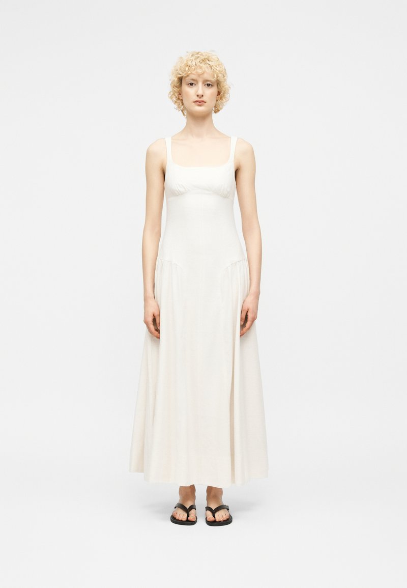 Νεαρό άτομο με κοντά σγουρά ξανθά μαλλιά, που φοράει μακρύ λευκό φόρεμα χωρίς μανίκια και μαύρα σανδάλια, στέκεται μπροστά σε ένα απλό φόντο.