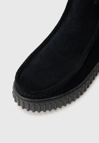 Bottine en suede noire avec une semelle en caoutchouc épaisse et striée, et un bout arrondi lisse, ornée de coutures le long du bord.