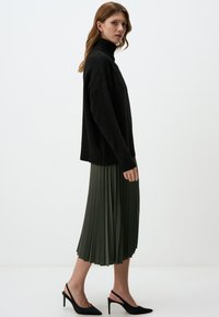 Schwarzer Strickpullover mit Rollkragen, kombiniert mit einem knielangen, plissierten olivgrünen Rock und schwarzen Slingback-Absätzen vor einem schlichten Hintergrund.