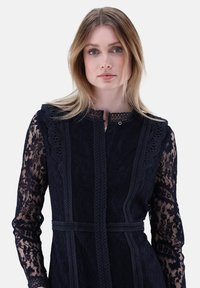 Navy-blaues Spitzenkleid mit langen Ärmeln, aufwendigen Designs, Knopfverschluss und einer taillierten Silhouette mit einem Stoffgürtel. Strukturierte und detaillierte Musterung.