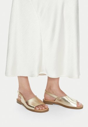 Sandalias metálicas doradas con punta abierta, correa trasera y suela plana. Textura suave con un diseño minimalista. Combinadas con un vestido blanco.