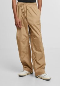 Beige, loose-fitting cargo pants made from durable fabric, featuring an elastic waistband with drawstring, side pockets, and minimal stitching details.