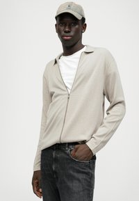 Lys beige zip-up jakke med krave, parret med en hvid T-shirt og mørke jeans. Modellen bærer en lys farvet kasket.