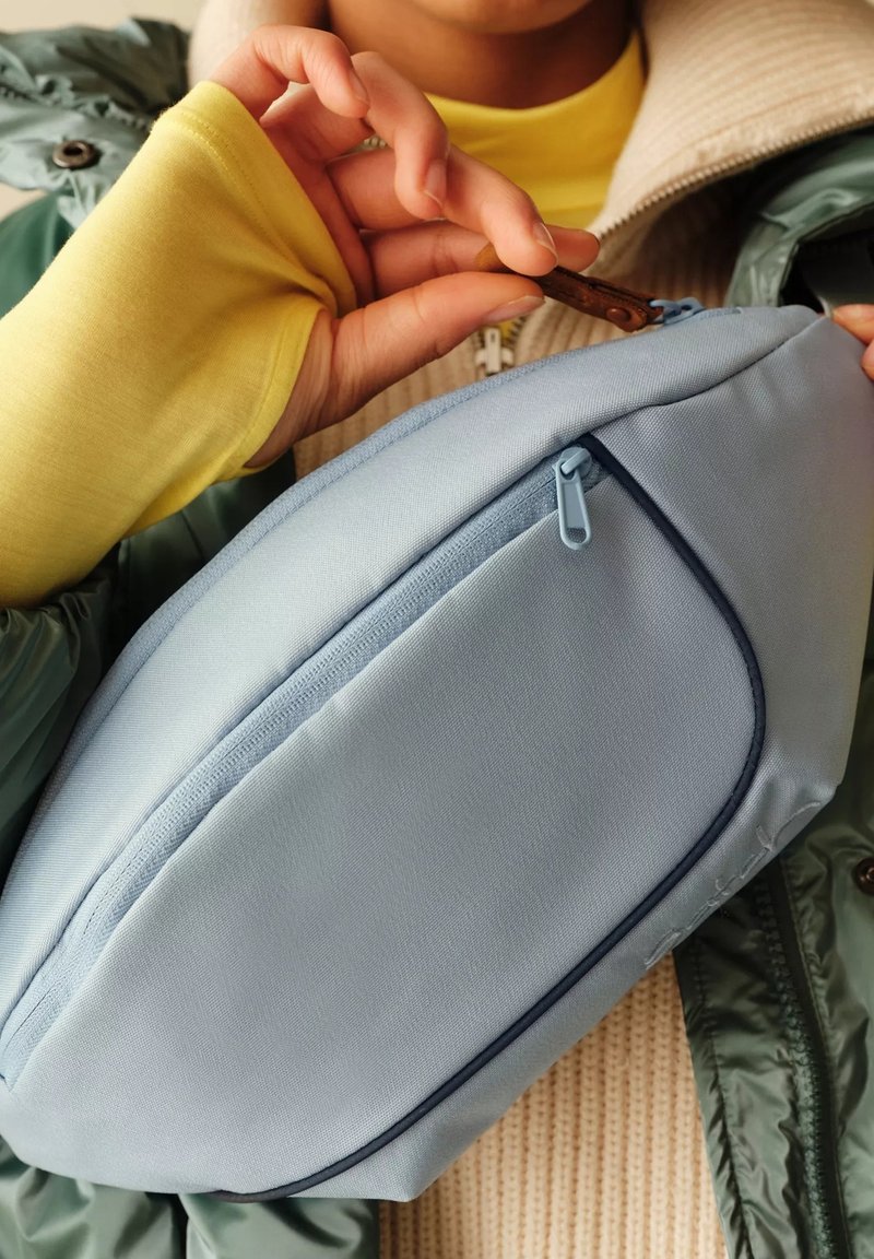 Hellblaues Stoff-Crossbody-Tasche mit einem Reißverschlussfach, runder Form und marineblauen Akzenten. Glatte Textur, gehalten von einer Hand in der Nähe einer Jacke.