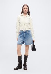 Γυναίκα που φοράει κρεμ ζακέτα με φερμουάρ, τζιν σορτς, ψηλές σκούρες καφέ μπότες, κρατώντας μικρή μαύρη τσάντα, στέκεται μπροστά σε απλό λευκό φόντο.