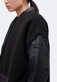 Femme aux cheveux foncés en queue de cheval portant une veste noire texturée avec des manches noires lisses et un petit patch sur le haut du bras.