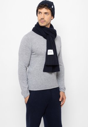 Donkerblauwe gebreide beanie en geribde sjaal, gecombineerd met een lichtgrijze aansluitende trui. De trui heeft lange mouwen, een eenvoudige stijl en een ronde hals.