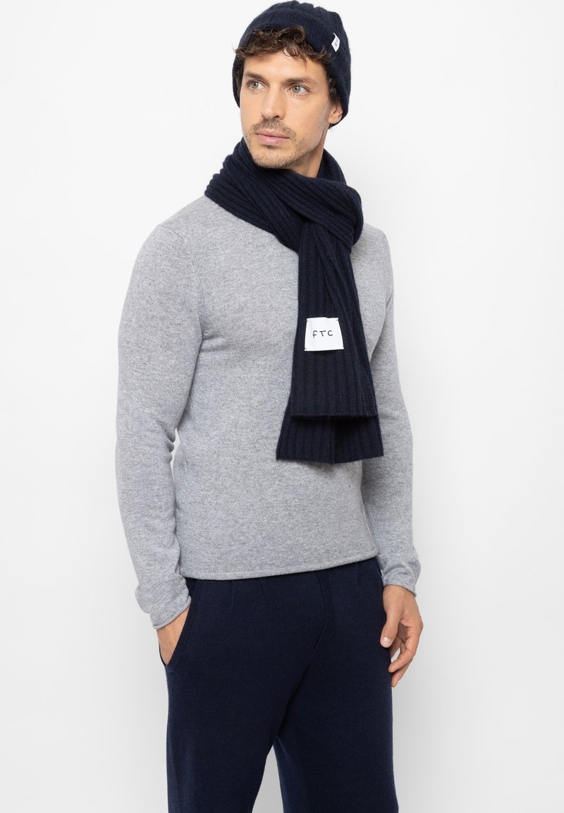 Donkerblauwe gebreide beanie en geribde sjaal, gecombineerd met een lichtgrijze aansluitende trui. De trui heeft lange mouwen, een eenvoudige stijl en een ronde hals.