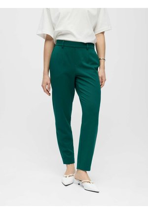 Femme portant un pantalon taille haute fuselé couleur sarcelle, un haut blanc à manches courtes, des chaussures blanches à talons bas pointues et un bracelet argenté sur fond blanc.