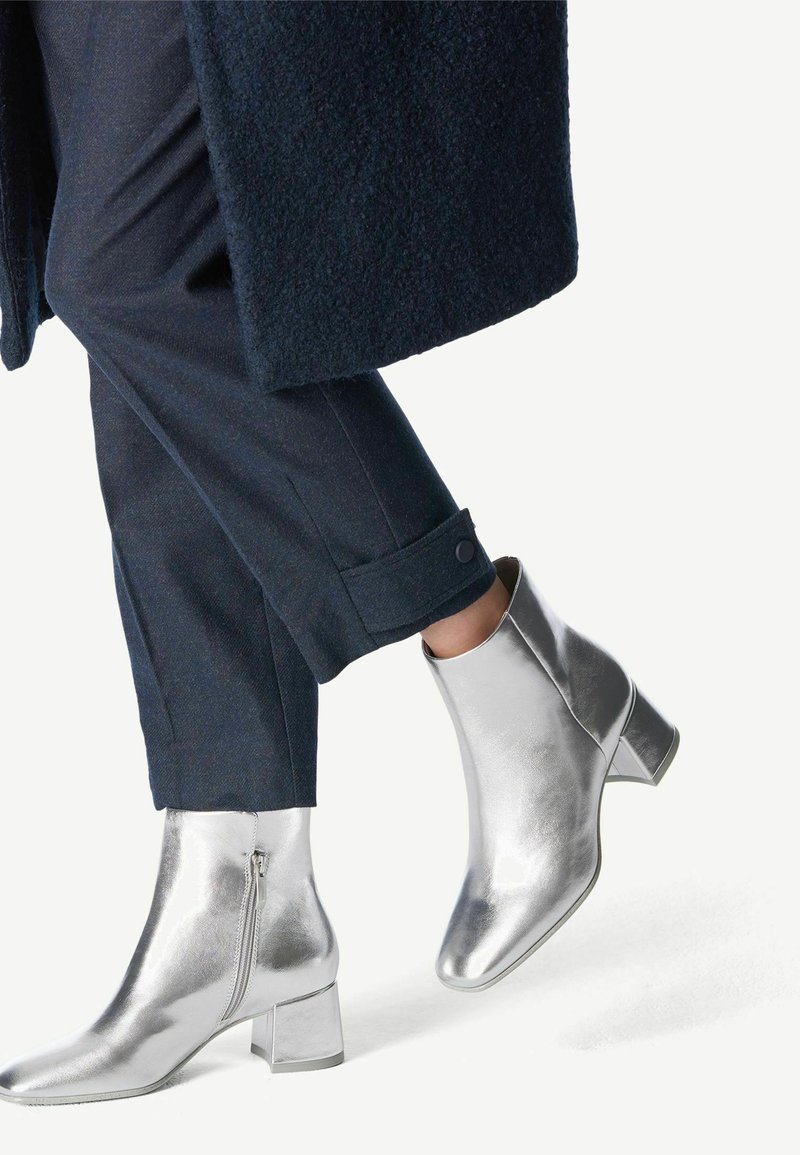 Metallic zilveren enkellaarzen met een vierkante neus, ritsdetails en een chunky hak, gecombineerd met marineblauwe omgeslagen broeken.
