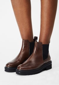 Botas de tobillo de cuero marrón con paneles elásticos laterales negros y suelas gruesas negras, llevadas sobre piernas desnudas frente a un fondo blanco.