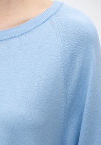 Pull en tricot bleu clair montrant l'encolure, la couture de l'épaule et le tissu à texture fine sur le haut du corps d'une personne.