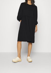 Black long-sleeve dress with a gathered waist and side pockets, paired with beige wooden clogs featuring a smooth finish and decorative studs.