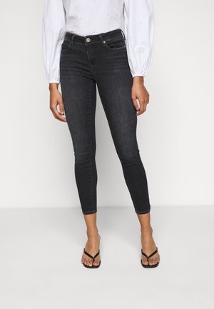 Frau trägt schwarze Skinny-Jeans mit hohem Bund, kombiniert mit einer weißen Bluse mit langen Ärmeln und schwarzen Riemchensandalen.
