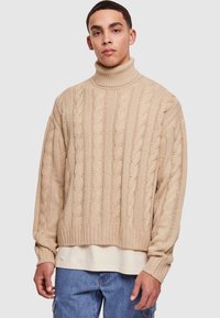 Pulover bej cu guler înalt din tricot cu model împletit, având manșete și tiv cu nervuri, cu o croială lejeră și un model texturat, purtat peste o cămașă crem.