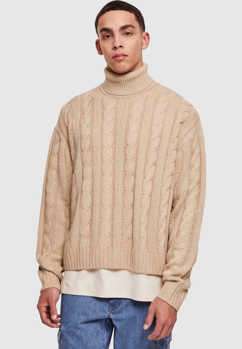 Pulover bej cu guler înalt din tricot cu model împletit, având manșete și tiv cu nervuri, cu o croială lejeră și un model texturat, purtat peste o cămașă crem.