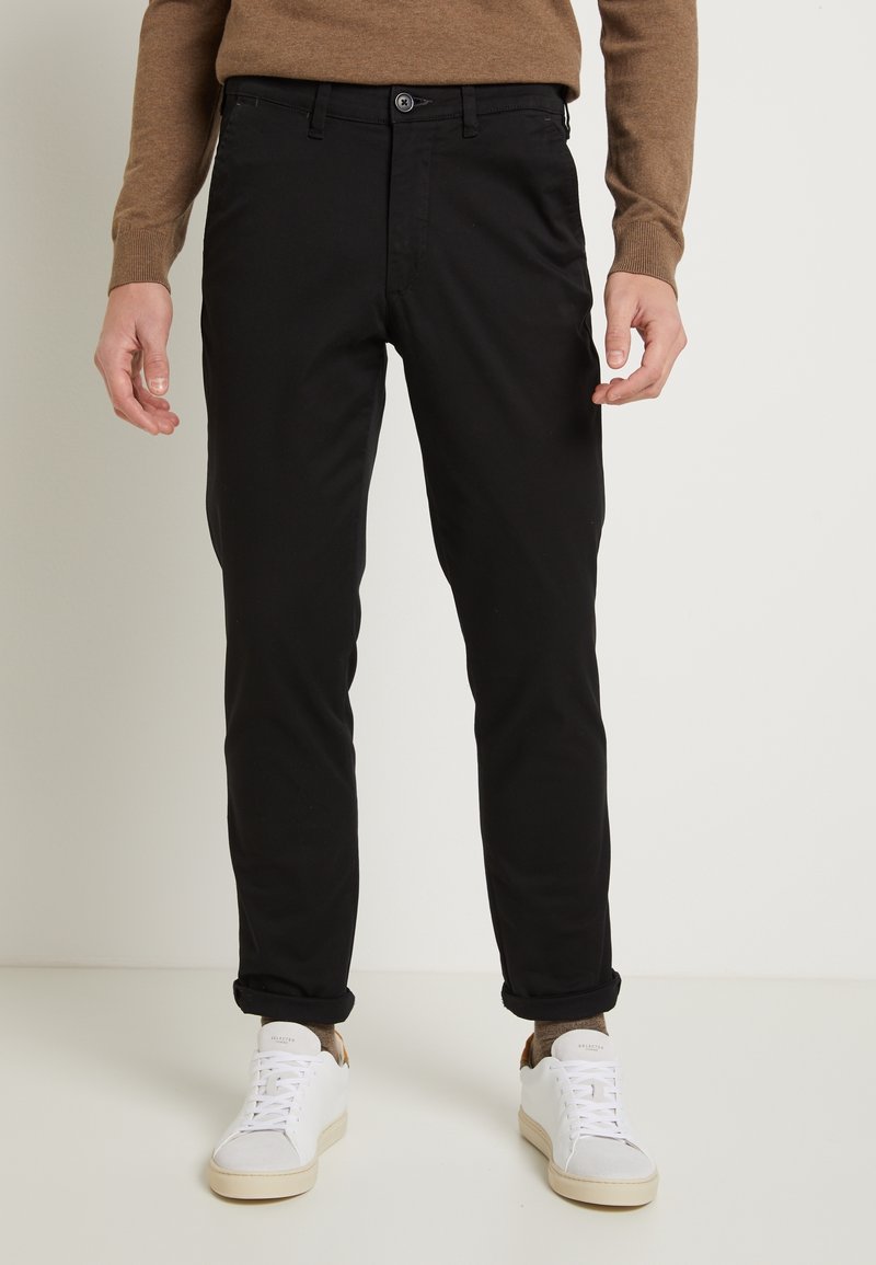 Black cotton chinos with a slim fit, featuring a button closure, pockets, and rolled cuffs, paired with white sneakers.