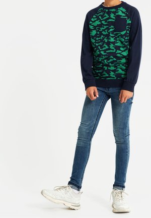 Kind draagt een marineblauwe camoufleretrui, strakke blauwe spijkerbroek en witte sneakers, staand met één voet licht opgetild.