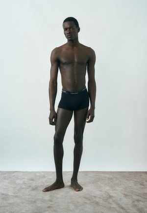 Νέος άνδρας στέκεται ξυπόλητος σε χαλί, φορώντας μαύρα μποξεράκια Calvin Klein πάνω σε μονόχρωμο ανοιχτό φόντο.