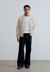 Άνδρας που στέκεται σε εσωτερικό χώρο φορώντας μια μπεζ ζακέτα με κουμπιά, λευκό μπλουζάκι, μαύρο παντελόνι με φαρδιά πόδια και καφέ παπούτσια, μπροστά σε έναν απλό λευκό τοίχο.