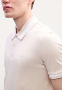 Cămașă polo bej deschis, realizată dintr-un material fin. Prezintă un guler cu dungi albe, placket cu două butoane și un logo brodat subtil „BOSS”.